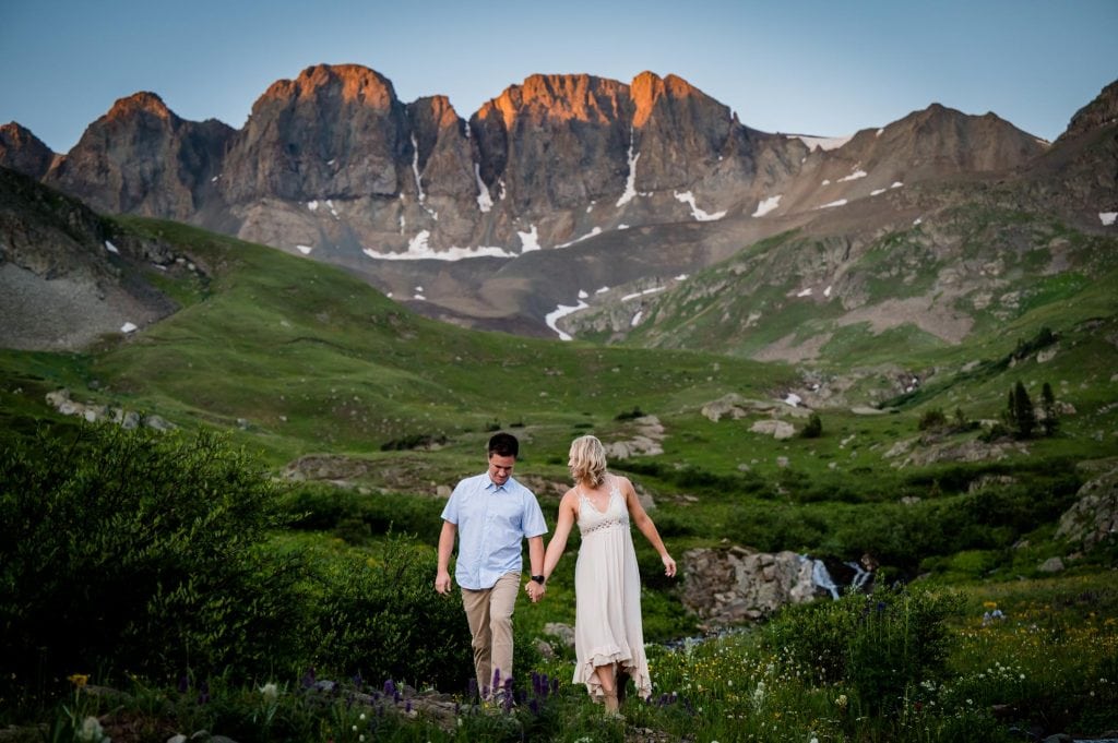 a couple walks hand in hand carefully around the wildflowers in American Basin at their sunrise adventure engagement session in Colorado