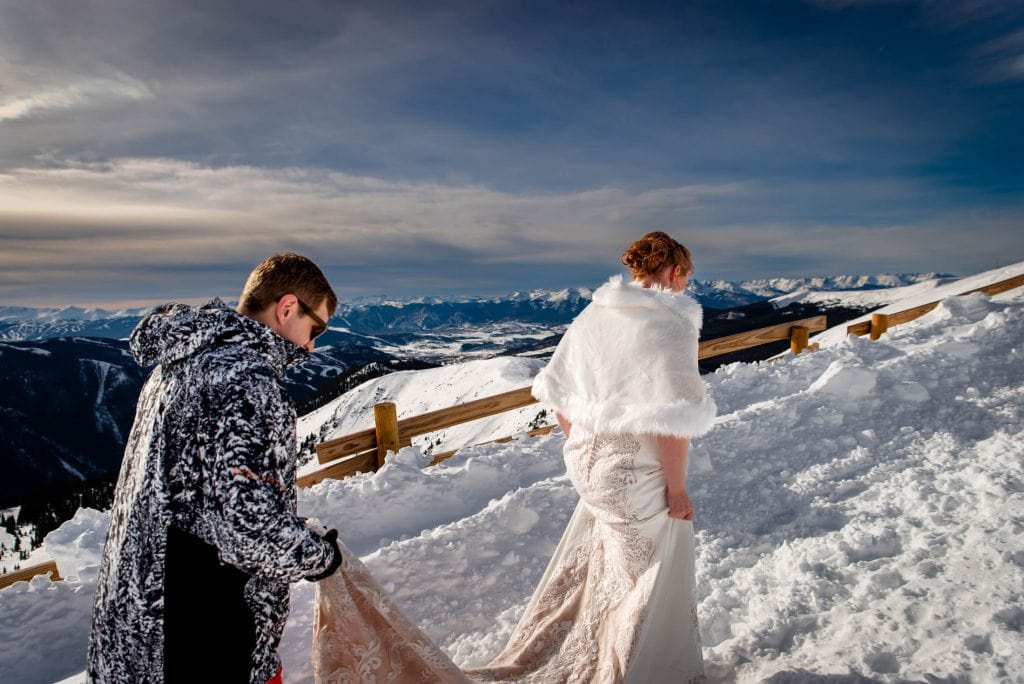 a groom helps his bride walk up the side of a mountain in her wedding dress for their snowboarding elopement at a-basin in colorado