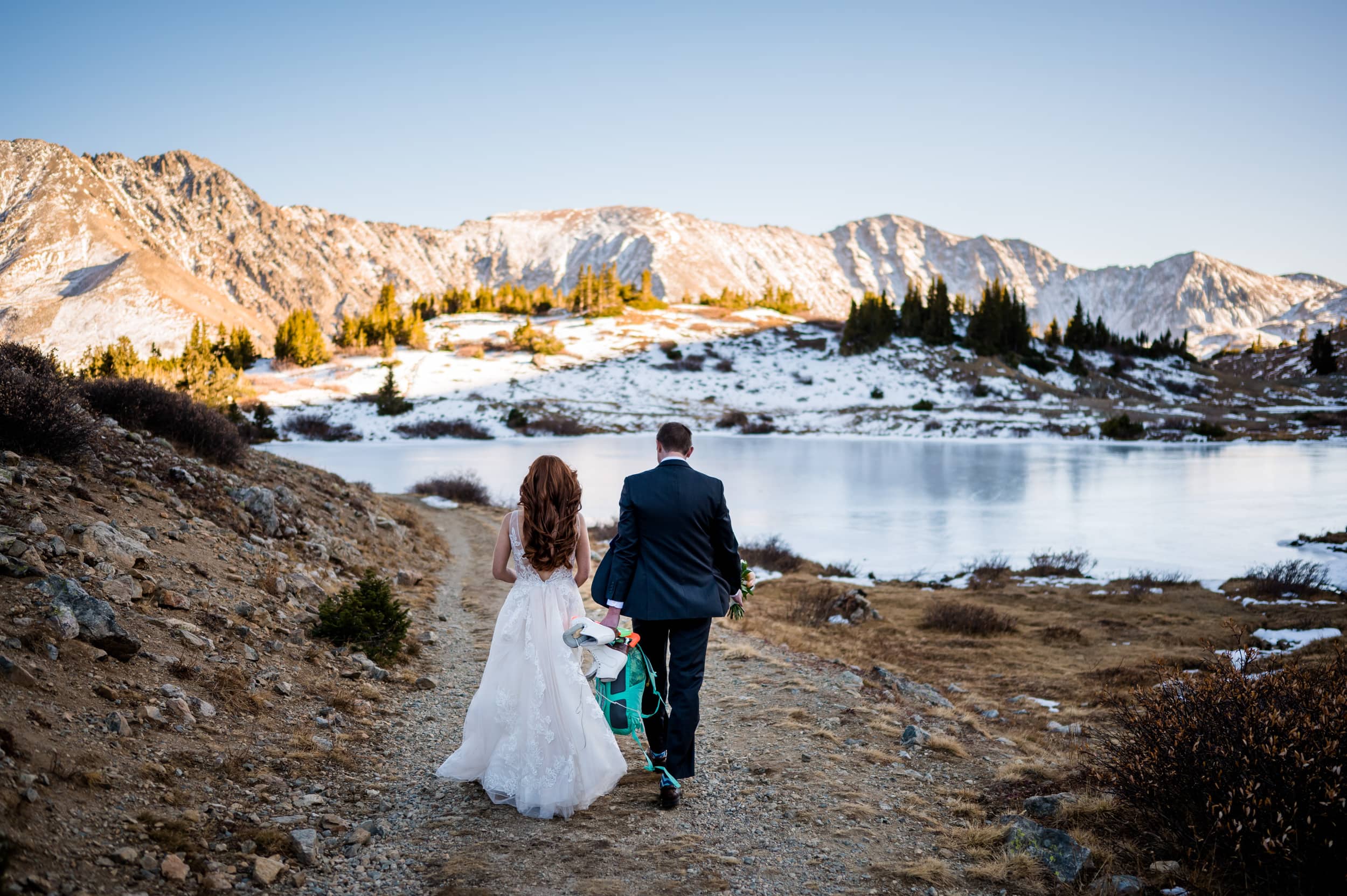 A marrying couple walks together in the Rocky Mountains towards a frozen alpine lake to ice skate on their elopement day