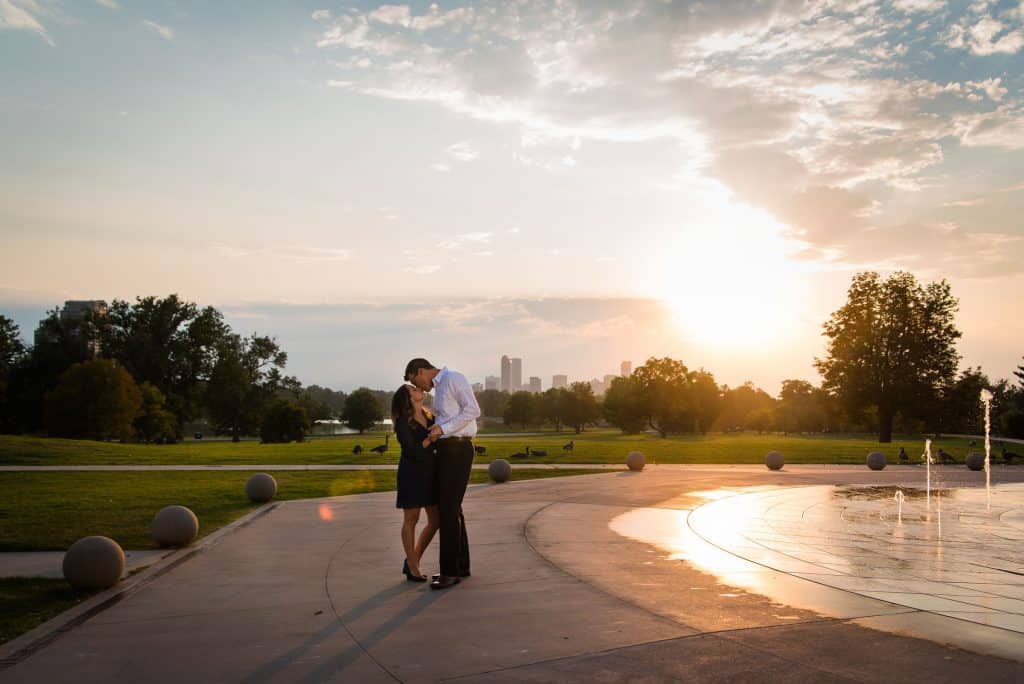 A couple kisses in the fountains at City park in Denver with the sunset falling behind the skyline and the mountain view.