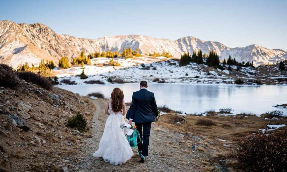 A groom holds his bride's ice skates as they walk towards a frozen backcountry alpine lake in the Arapahoe National Forest in Colorado