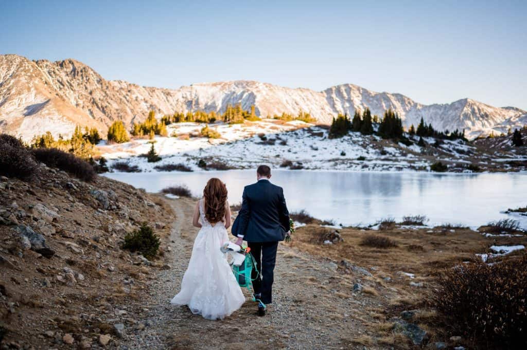 A groom holds his bride's ice skates as they walk towards a frozen backcountry alpine lake in the Arapahoe National Forest in Colorado