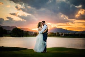 A couple embraces in front of a lake with mountains in the background during a stormy orange and grey sunset at the Barn at Raccoon Creek wedding venue in Colorado