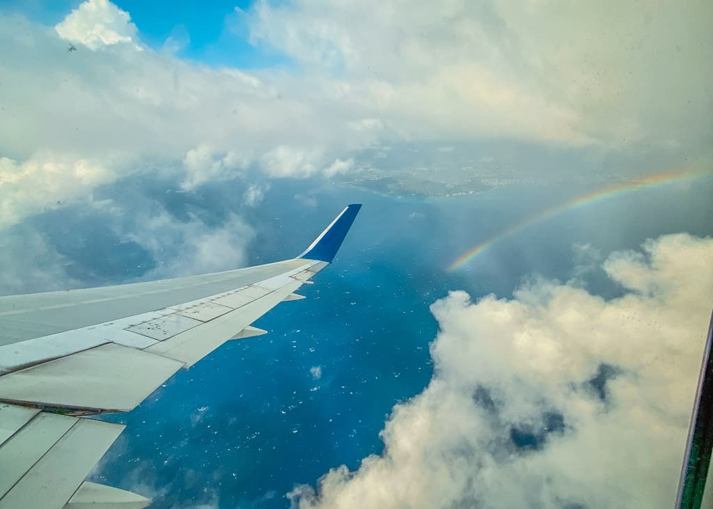 An airplane in the clouds flying over Hawaii with a rainbow coming out of the ocean