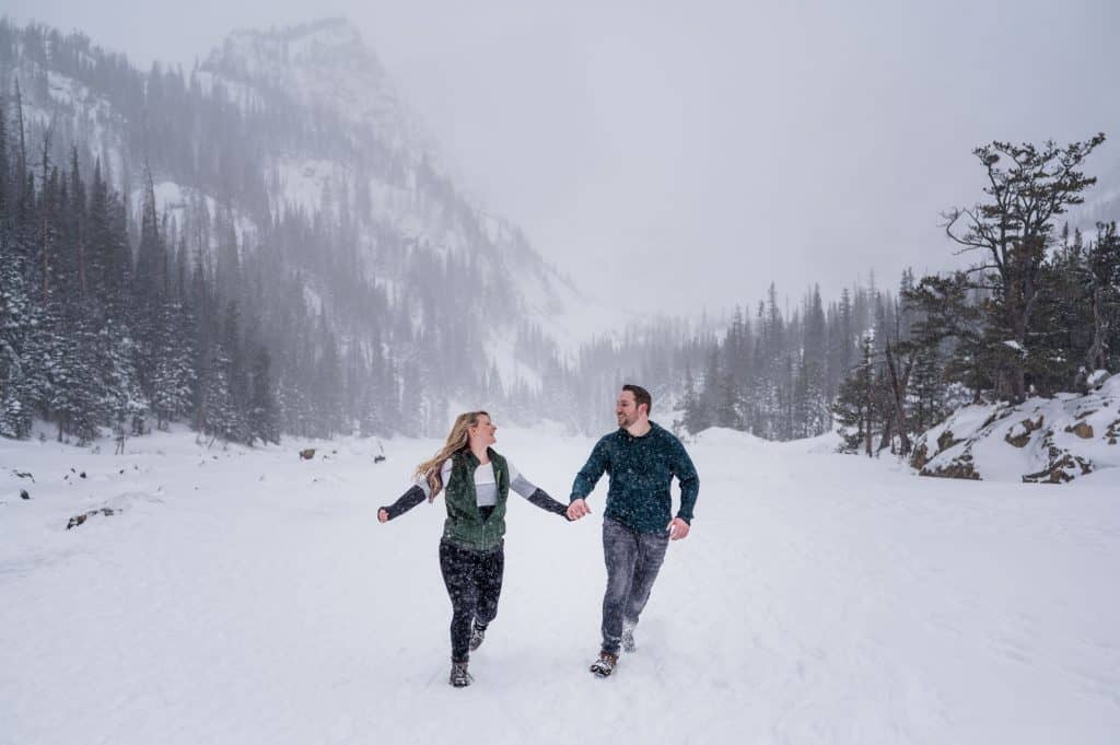A couple runs through a blizzard on a frozen lake in RMNP for their engagement photos