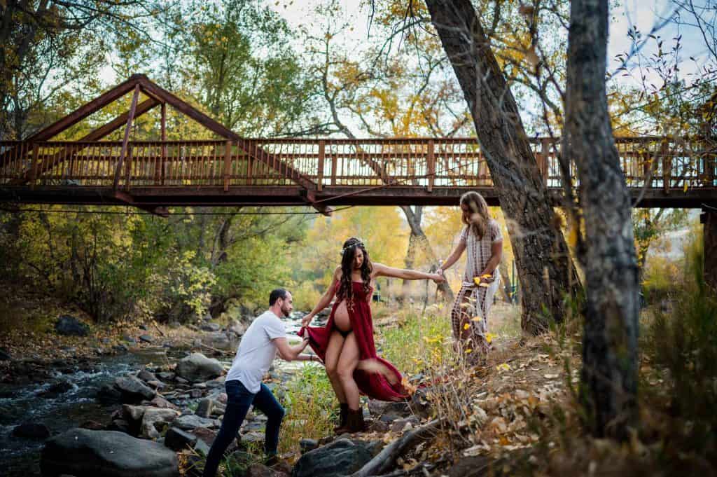 a throuple helps each other out over a creek for their maternity photos in Denver