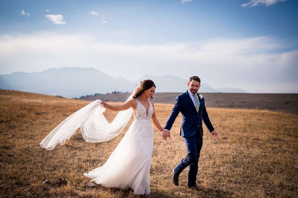 a bride and groom walk hand in hand through a grassy field with views of the Flatirons in Boulder, Colorado on their wedding day