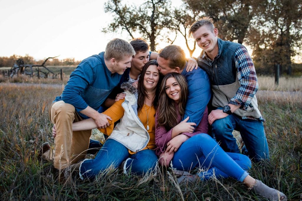 Older teenage children all snuggle mom in the grass while dad gives her a forehead kiss at their family photography session in Colorado