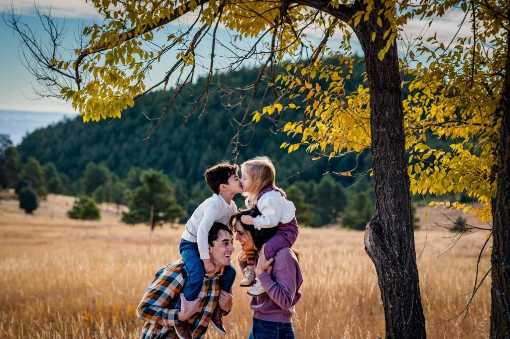 Toddler siblings are on parents' shoulders and giving a kiss to each other during at their outdoor mountain family adventure 