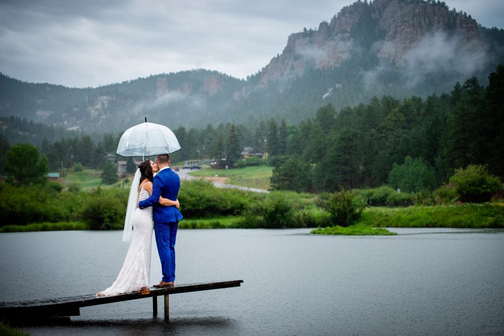 a couple stands on a dock under an umbrella as it rains in the mountains after their wedding ceremony at wedgewood mountain view ranch in Colorado