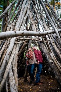 a couple is back to back and hand in hand trying kiss over their shoulders in a teepee in Evergreen, Colorado for their adventure engagement photography
