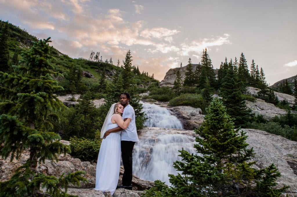 a bride warms her hands on her husband at their elopement near a waterfall in the mountains at sunset in Colorado