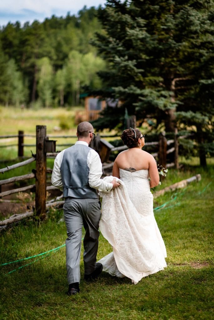 a groom holds his bride's dress as they walk in the mountains after their wedding ceremony at Meadow Creek Lodge and Event Center in Colorado