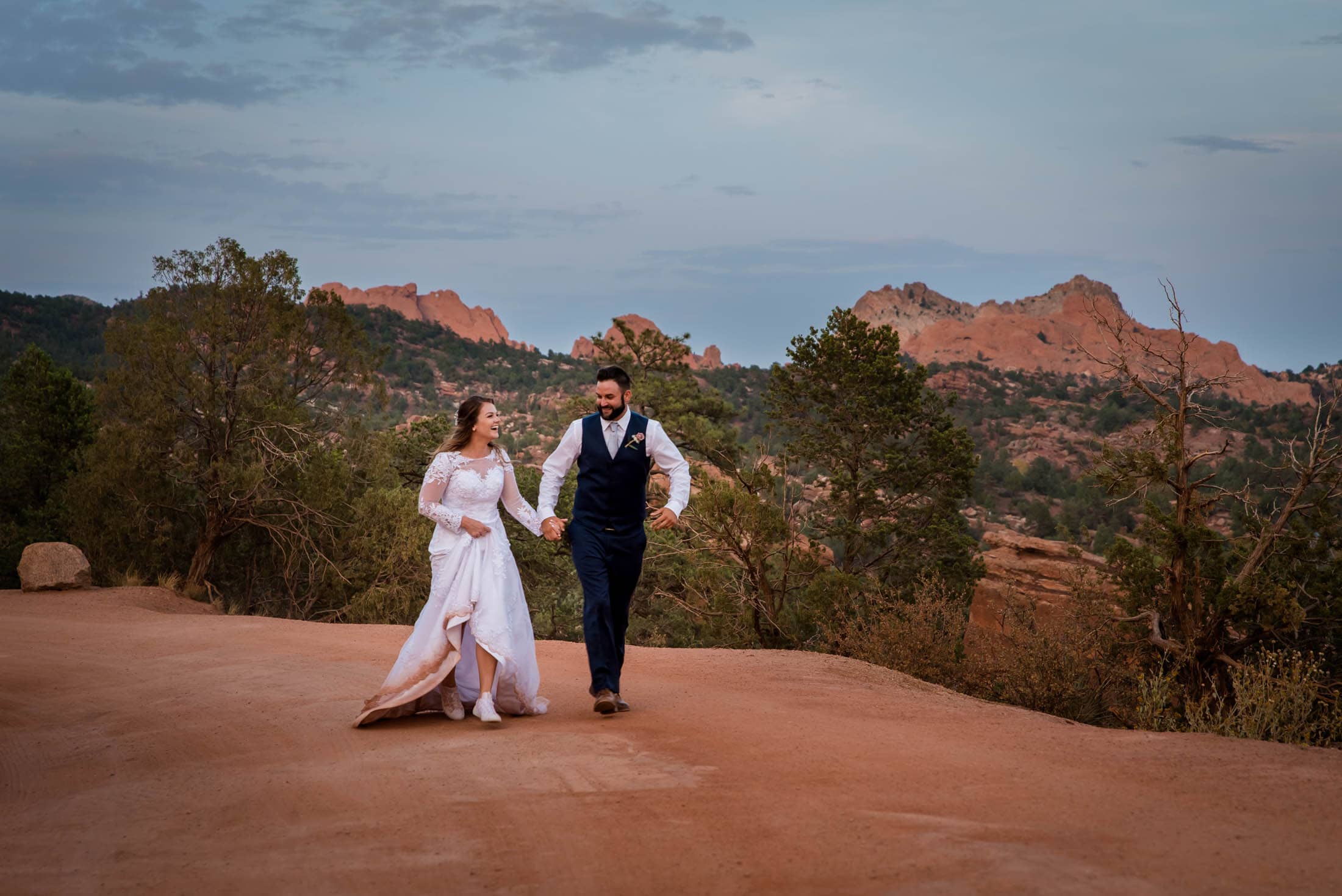 a couple runs hand in hand in the red dirt desert terrain at their Garden of the Gods Adventure Elopement