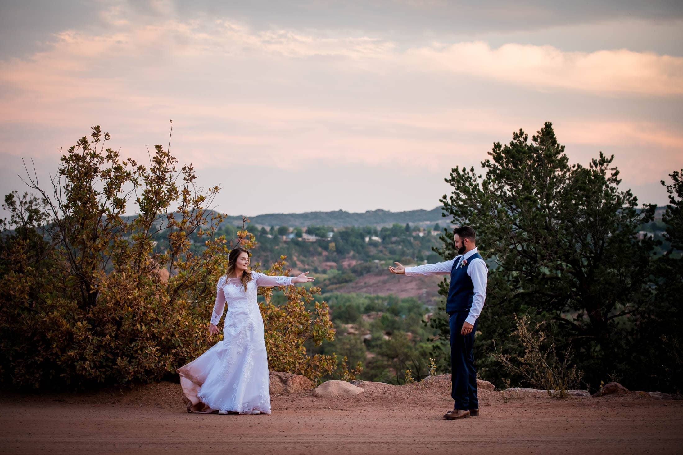A marrying couple reach for each other against a rusty red dirt backdrop at sunset near Garden of the Gods Colorado at their elopement
