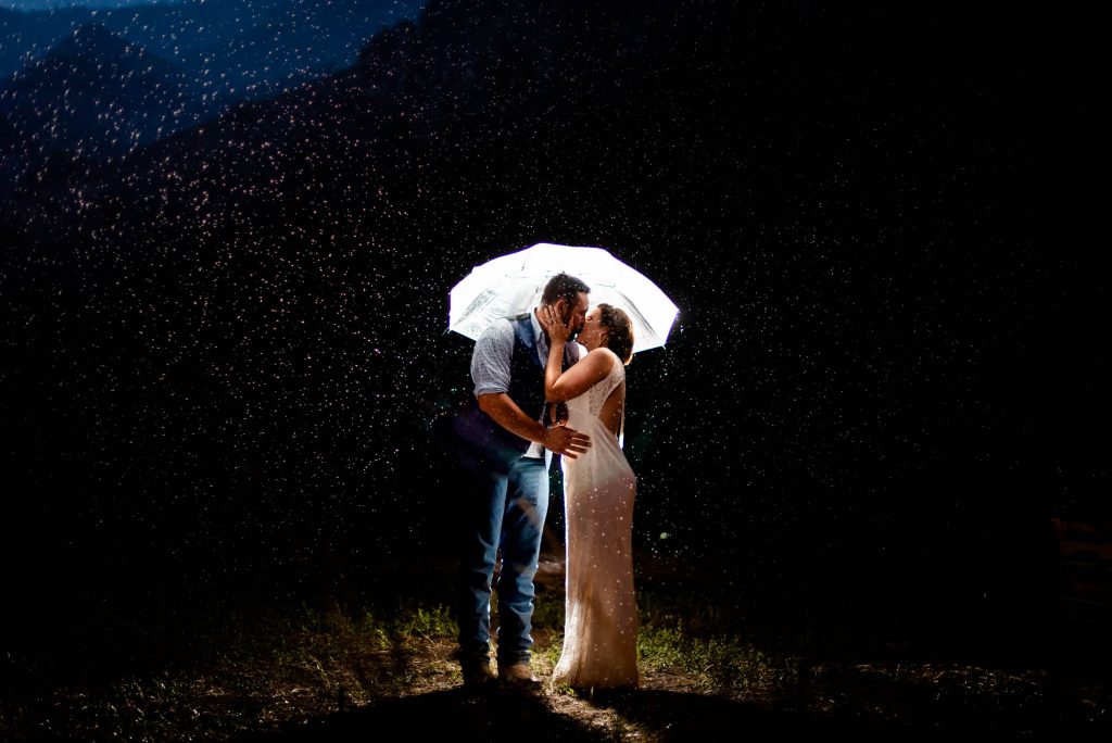 a couple kisses under a clear umbrella lit up by a light at night after their mountain wedding