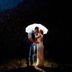 a couple kisses under a clear umbrella lit up by a light at night after their mountain wedding