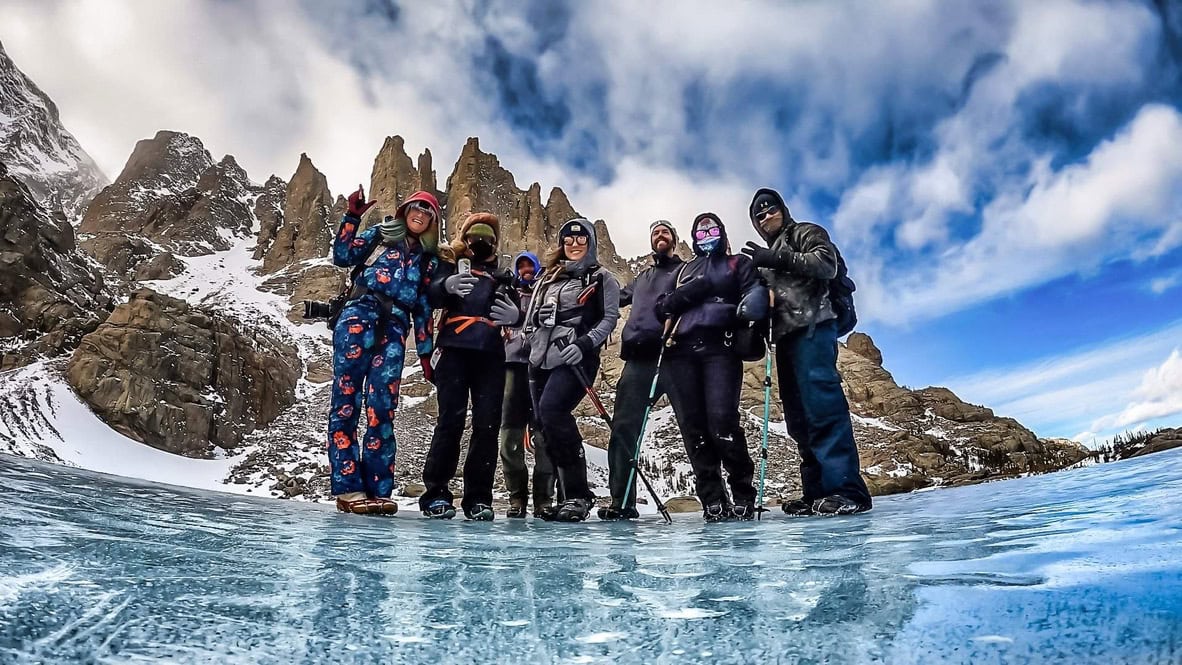 Friends stand on a frozen Sky Pond in RMNP in front of the spires after hiking up in the winter together. 